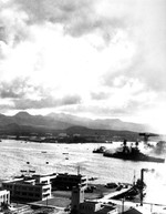 View from the Ford Island water tower across the Pearl Harbor turning basin during the Japanese air attack on 7 Dec 1941. Note the Hammerhead Crane and USS Nevada slipping by close to the Ten-Ten Dock.