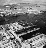 Aerial view of shipping in the Pearl Harbor Navy Yard repair basin with Ford Island beyond, 12 Dec 1943. Note the camouflage patterns painted on the roofs of the buildings.