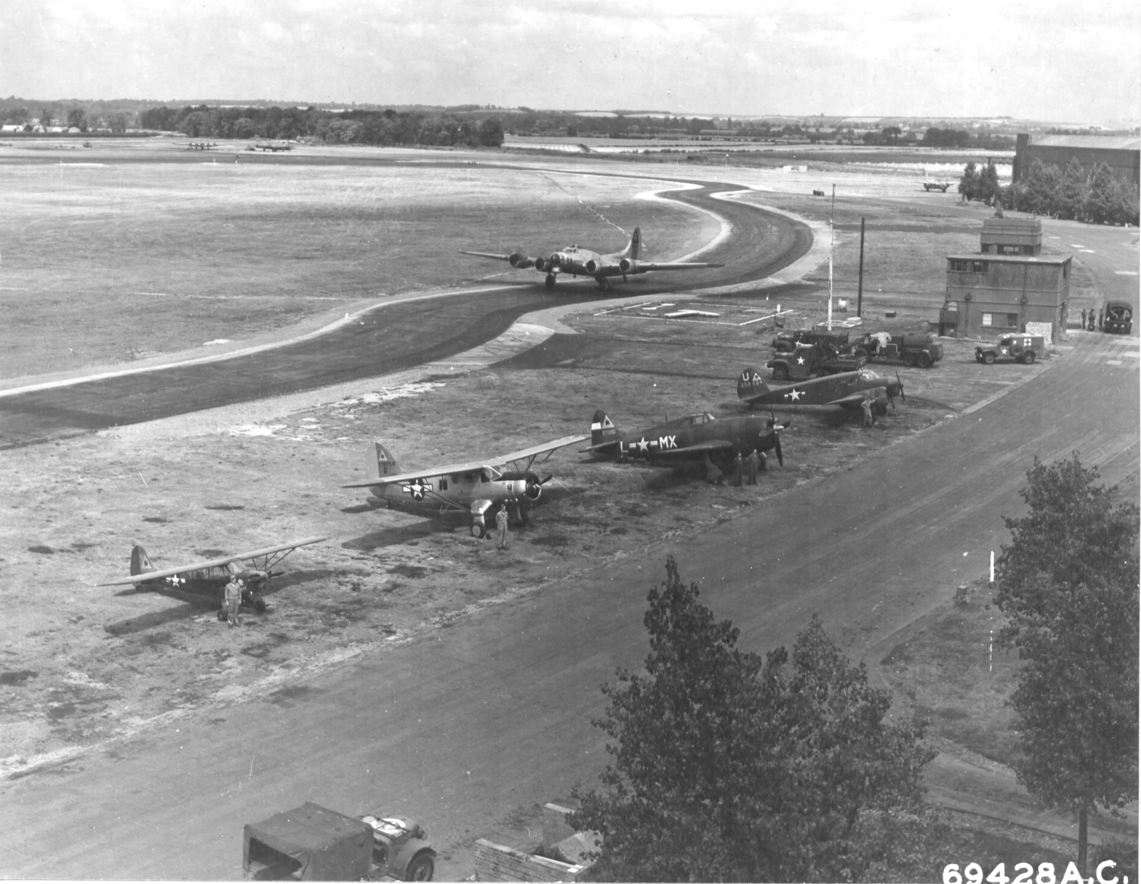 View of the control tower at RAF Bassingbourn, Cambridgeshire, England, UK, 1945. Note line of 91st Bomb Group “hack” aircraft: Piper L-4 Grasshopper, Noordyne O-64 Norseman, P-47 Thunderbolt, and Cessna UC-78 Bobcat.