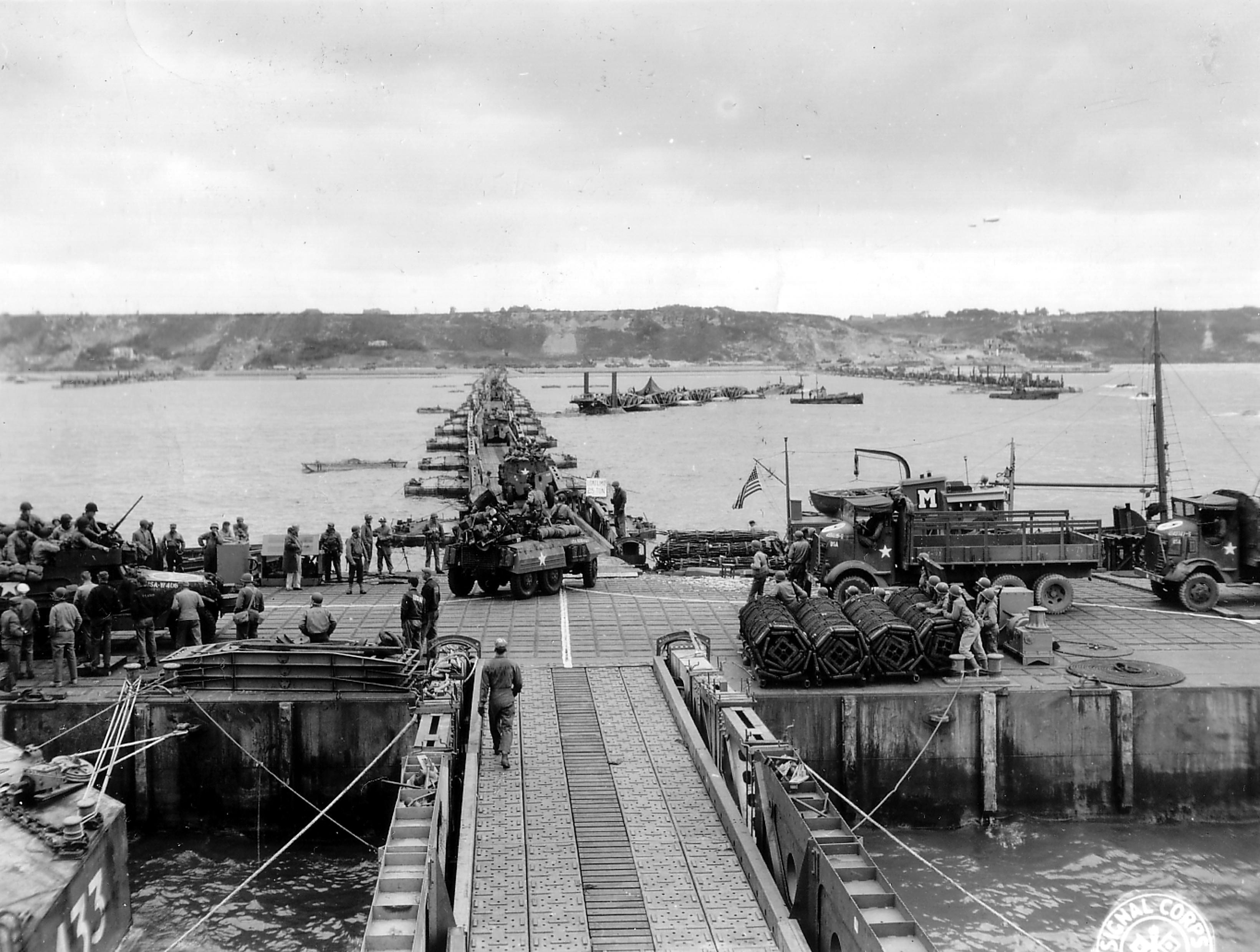 Military vehicles move ashore from Mulberry Artificial Harbor A, across a pontoon bridge, to Omaha Beach, Normandy, France, June 16 1944 (D+10). Visible are M3 Halftracks, an M8 Greyhound, and AFKWX 2½-ton Trucks.