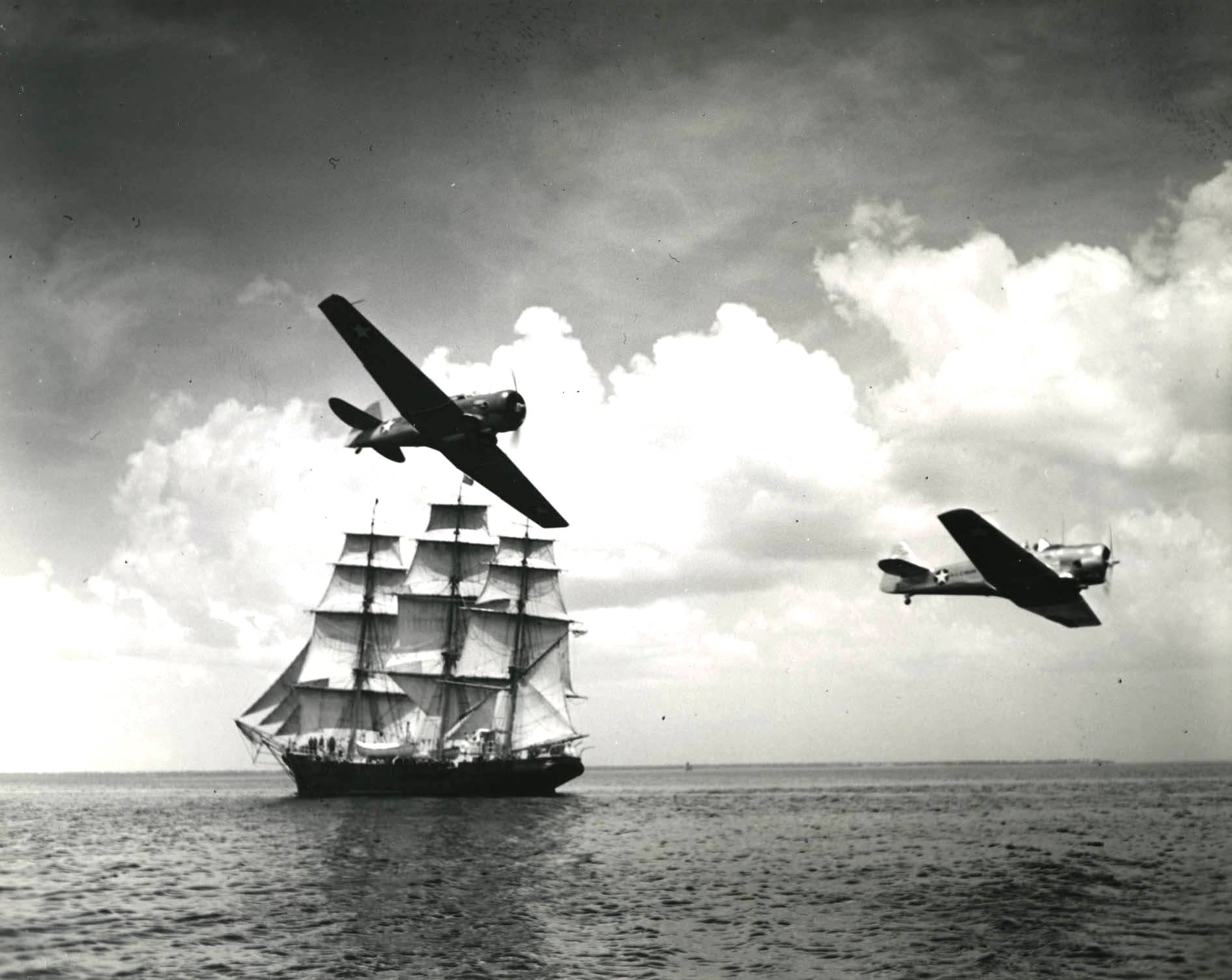 US Navy SNJ Texan training aircraft making a low level pass near a three-masted sailing ship, probably along the east coast of the United States, 1942
