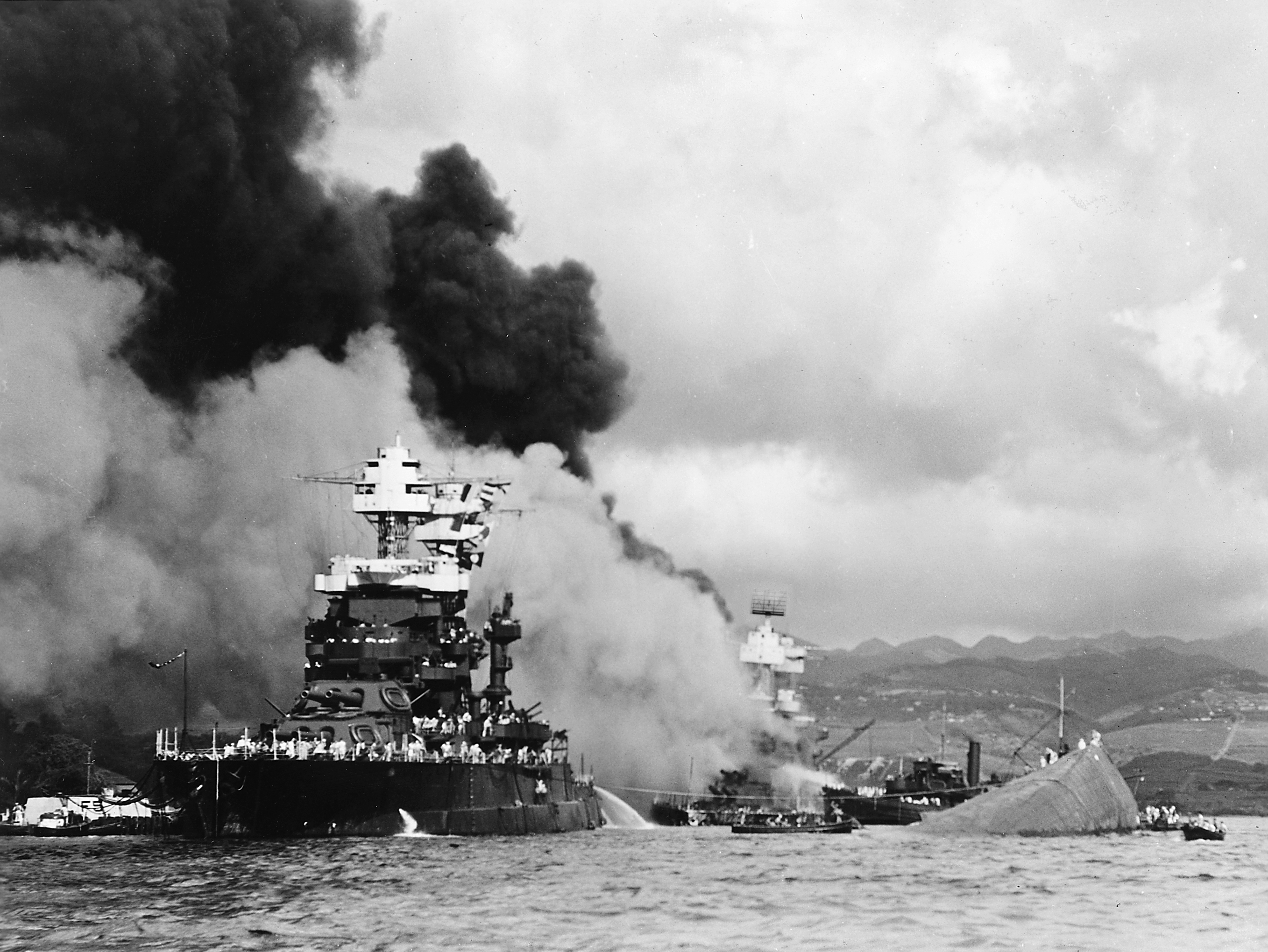 A bow-on view of the comparatively lightly damaged battleship USS Maryland with the burning USS West Virginia behind her and the capsized USS Oklahoma beside her, Pearl Harbor, US Territory of Hawaii, 7 Dec 1941