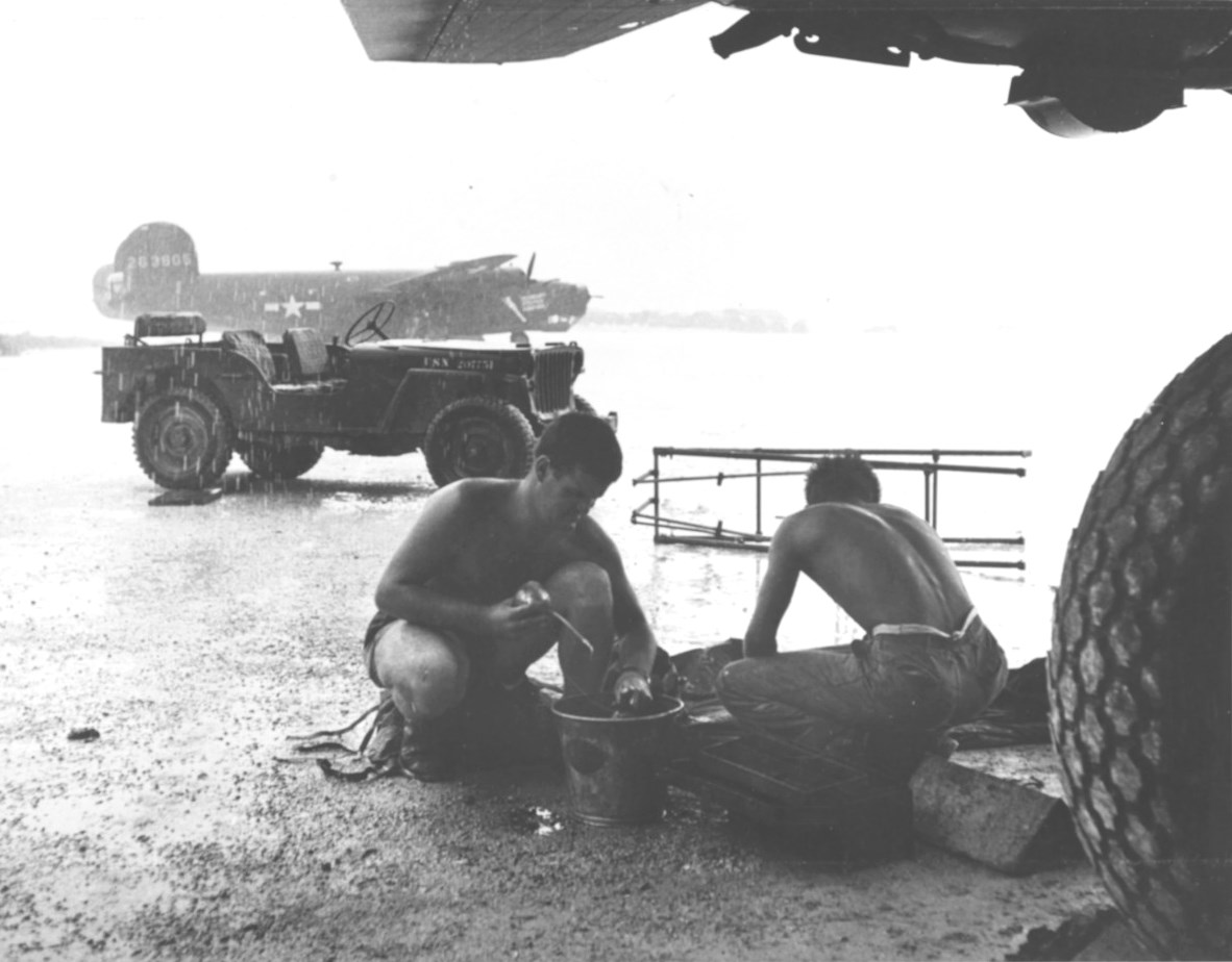 US Navy maintenance crews with Bombing Squadron VB-106 clean engine parts under an airplane wing during a downpour at Momote Airstrip, Los Negros, Admiralty Islands, Mar 1944. Note Jeep and Army SB-24D Liberator.