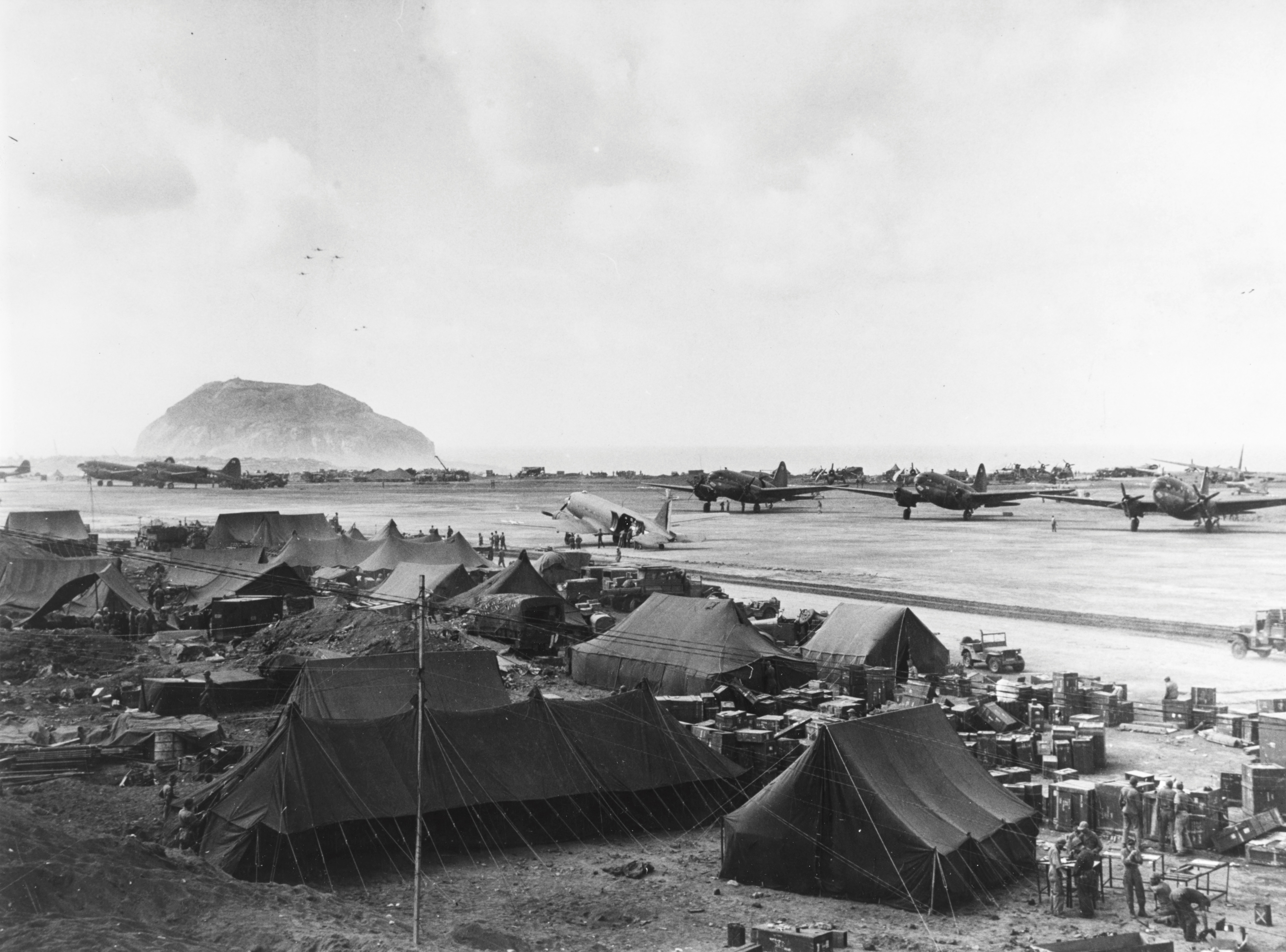 A Douglas R4D (C-47) transport plane (center) loading wounded Marines on Iwo Jima's South Field bound for a Navy hospital on Guam, Mar 1945. Note Mount Suribachi to the left.