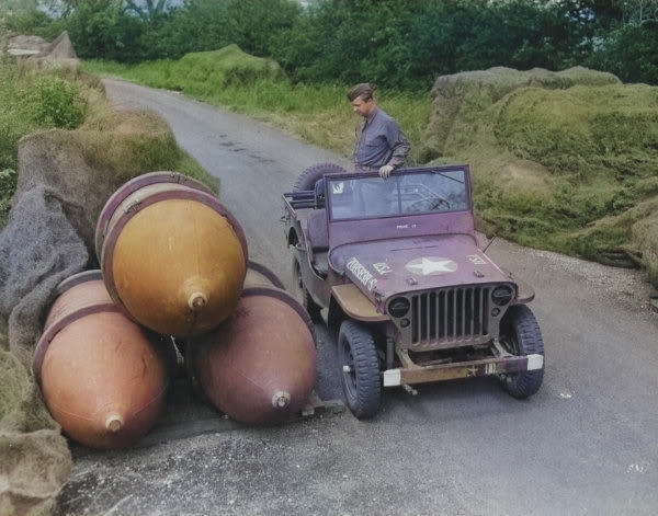 A member of the 2107th Ordinance-Ammo Battalion inspecting a store of 4000-pound bombs, some under camouflage netting, along the roadside at the Sharnbrook Ordinance  Depot, Bedfordshire, England, UK.  July 1943 [Colorized by WW2DB]