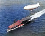 USS Langley underway off Cape Henry, Virginia, United States, 6 Oct 1943; note SNJ aircraft on flight deck and K-class airship from Lighter Than Air Squadron ZP-14 overhead.