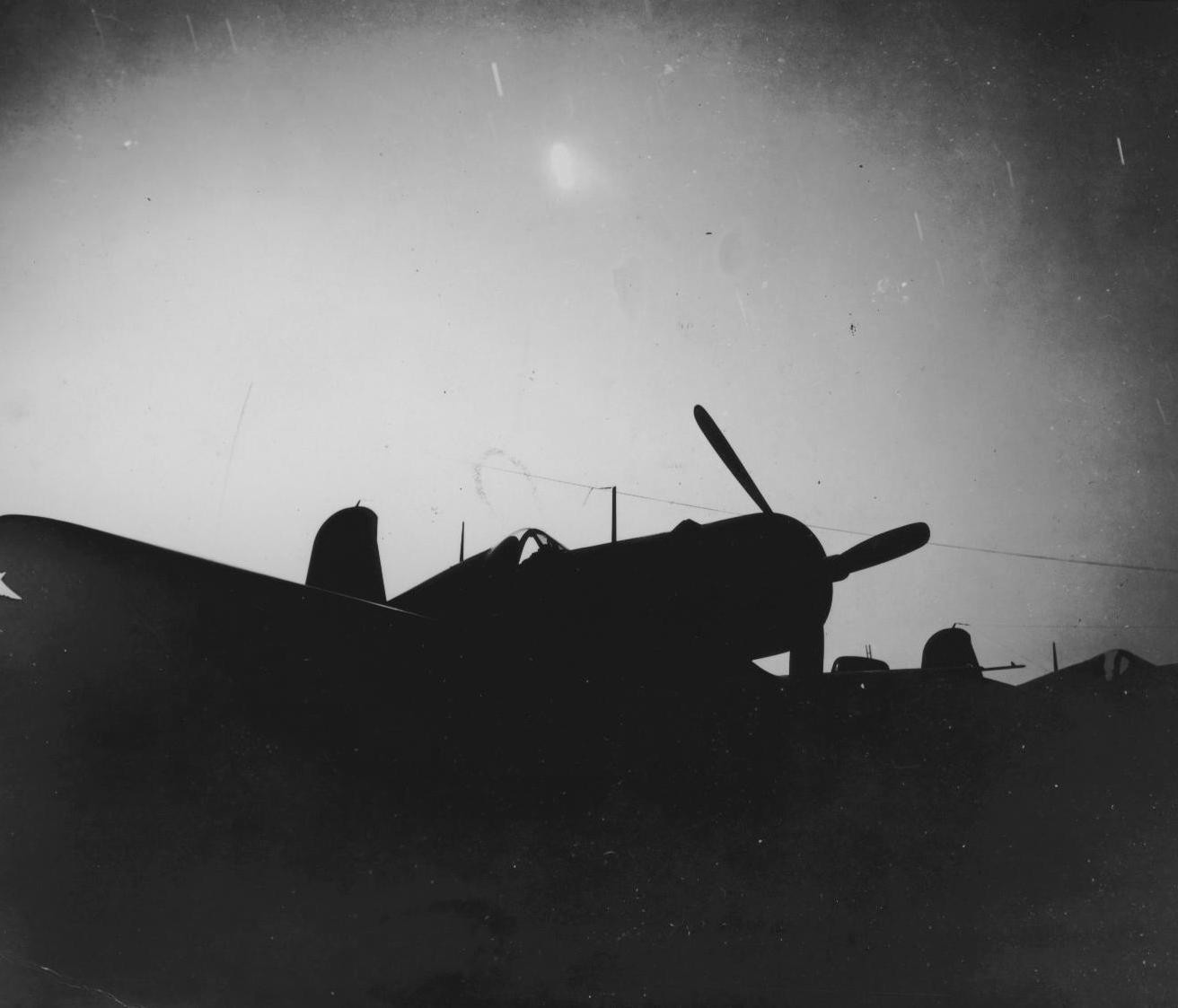 Silhouette of a USMC Corsair fighter seen against a burning supply dump, Yontan Airfield, Okinawa, Japan, Apr 1945