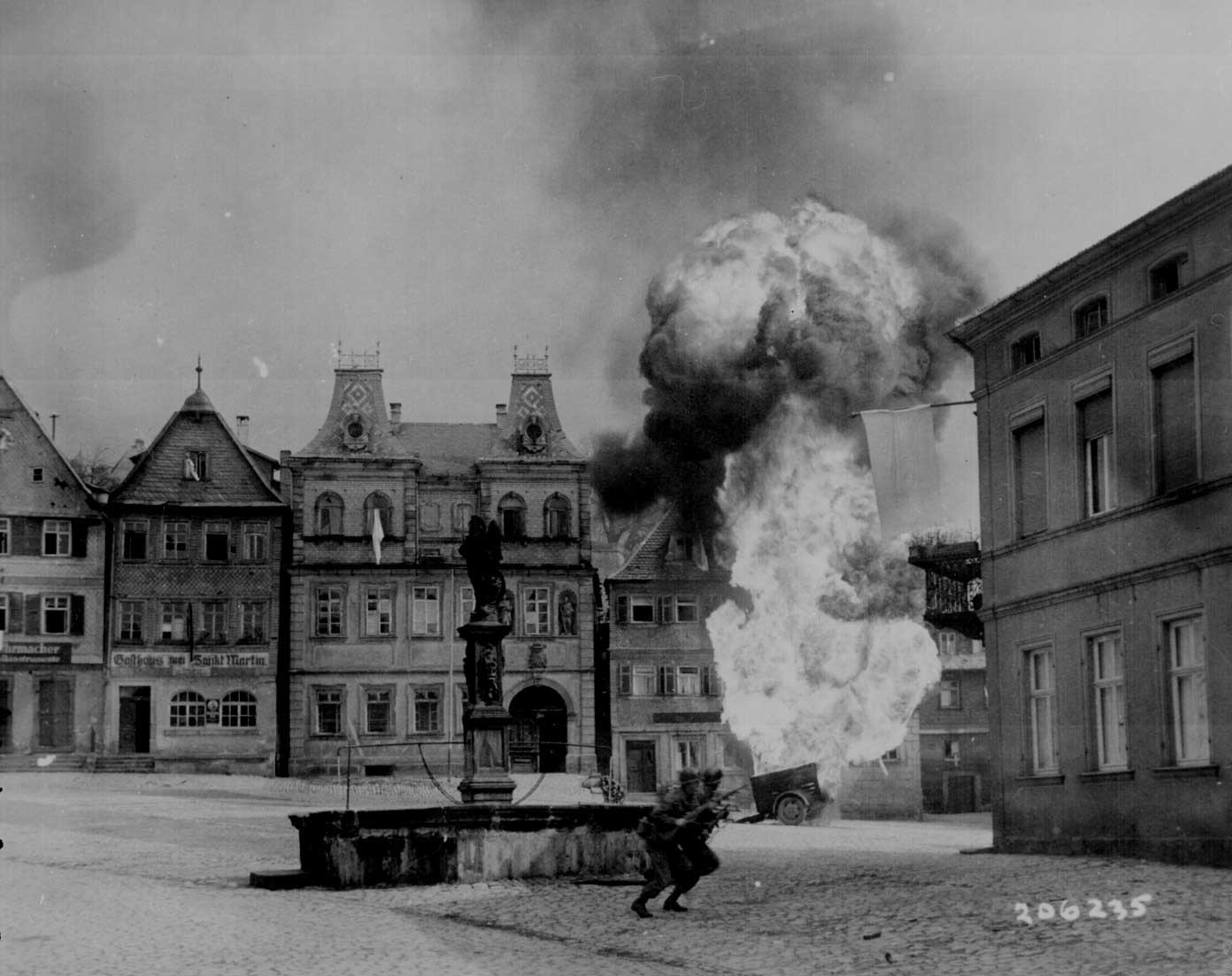 Men of US 101st Infantry Regiment running past a burning fuel trailer in square of Kronach, Bayreuth, Germany, 14 Apr 1945