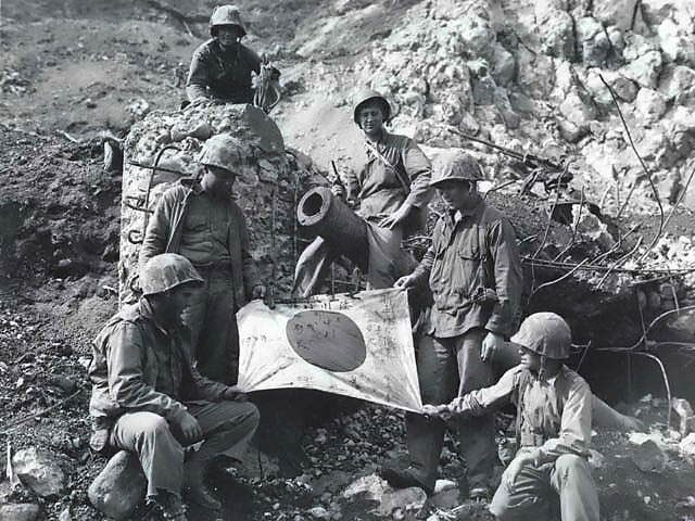 US Marines PFC. J. L. Hudson, Pvt. K. L. Lofter, PFC. Paul V. Parces, Pvt. Fred Sizemore, PFC. Henrey Noviech, and Pvt. Richard N. Pearson posing with a captured Japanese flag, Iwo Jima, Feb 1945