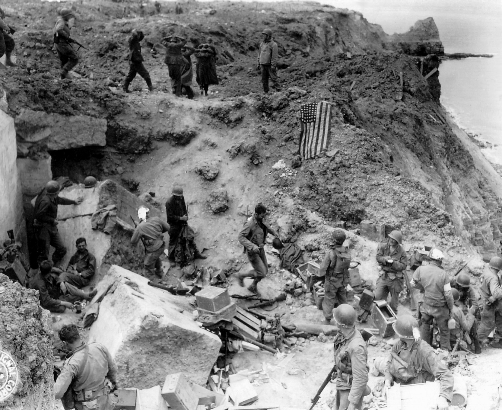 German prisoners being taken away atop a cliff at Pointe du Hoc, Normandy, France, 8 Jun 1944; note American flag draped on cliff wall to prevent friendly fire
