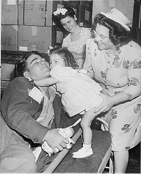 American Private First Class Lee Harper, wounded at Normandy, was greeted by his 2-year-old sister Janet, whom he had never seen before, as his mother and wife looked on, New York City, 1 Aug 1945