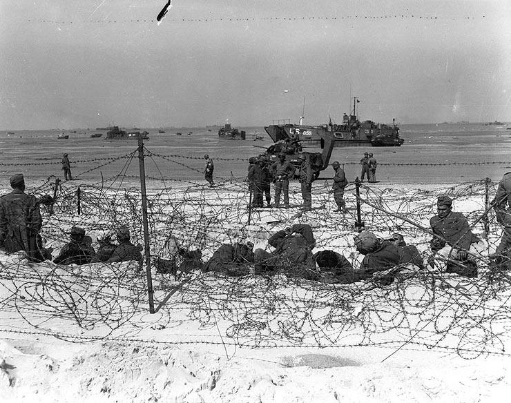 German prisoners of war in a barbed-wire enclosure on Utah Beach, Normandy, 6 Jun 1944; note M4 Sherman tank 'Delphia' and stranded LCT-855 in background