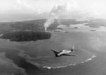 Smoke rising from Colonia Harbor on Yap, Caroline Islands as a TBM Avenger from the carrier Yorktown (Essex-class) flies overhead, 25-28 July 1944