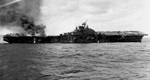 The carrier Franklin burning and listing badly after bomb hits aft set off more bombs and fueled aircraft, 19 Mar 1945. Note burning fuel pouring off the hangar deck and dislodged forward elevator.