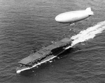 USS Langley underway off Cape Henry, Virginia, United States, 6 Oct 1943; note SNJ aircraft on flight deck and K-class airship from Lighter Than Air Squadron ZP-14 overhead.