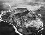 Aerial view of Mount Suribachi on Iwo Jima, Bonin Islands with the relatively flat island plain beyond, Feb or Mar 1945 as the battle raged on. Note landing ships beached at right.