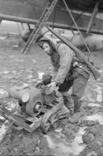 Corporal Lawrie Lever of RAF Mobile Repair Unit warming his hands with an air compressor which had just been used to service the engines of the Harrow aircraft in the background, Advanced Landing Ground B-56 Evere, Brussels, Belgium, 1945