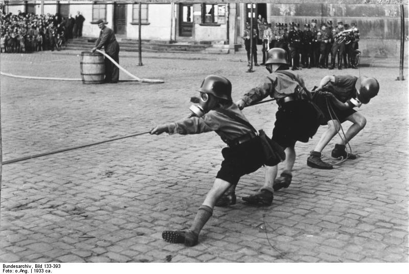 Hitler Youth members playing tug of war while donning helmets and gas masks, 1933