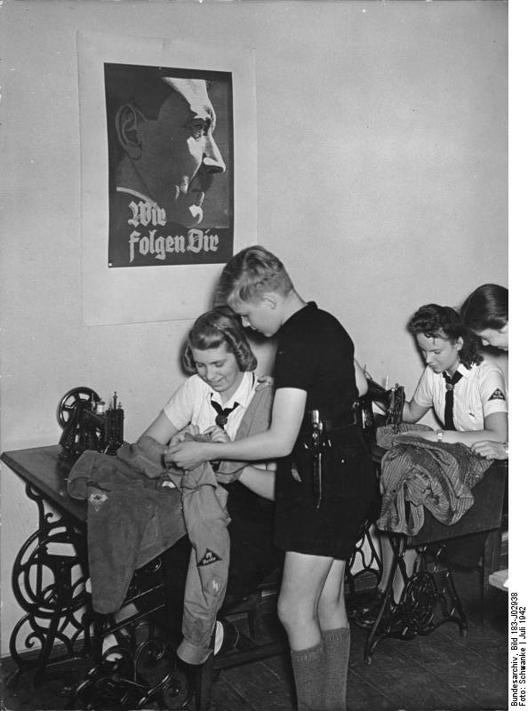 Members of the League of German Girls sewing in Berlin, Germany, Jul 1942; note boy with dagger and poster of Hitler 'We Follow You' on wall